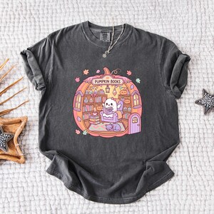 May include: Dark gray t-shirt featuring a colorful illustration of a pumpkin-shaped room with the text "Pumpkin Books". Inside, a ghost-like character reads a book, surrounded by bookshelves, a cat, and autumn leaves.