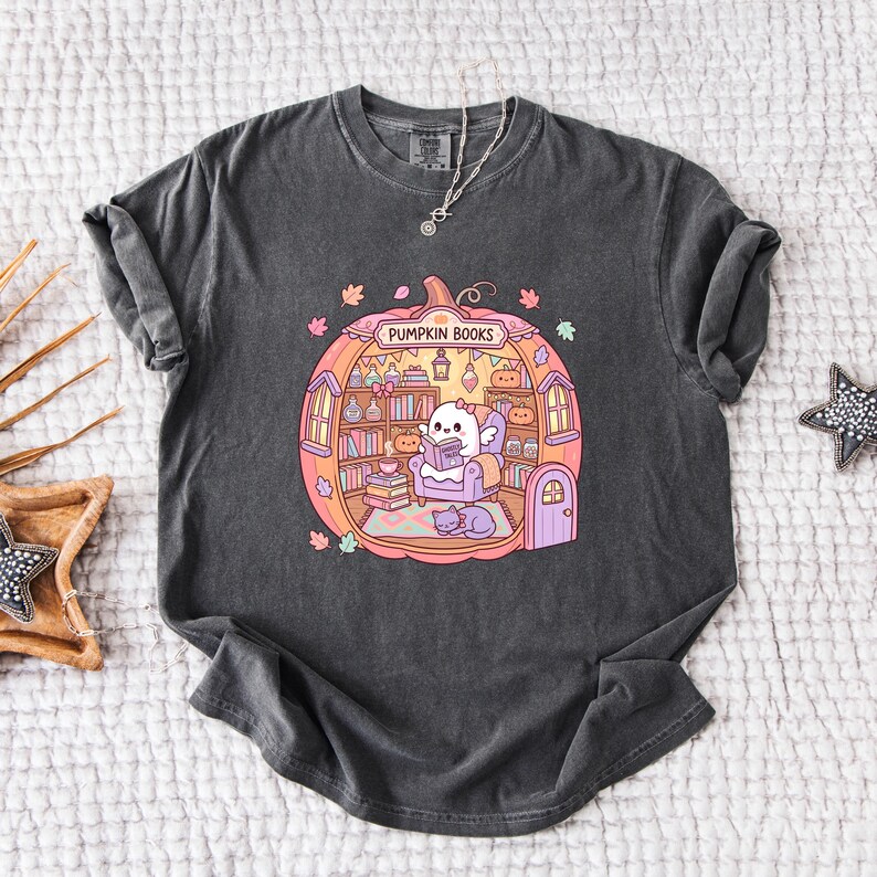 May include: Dark gray t-shirt featuring a colorful illustration of a pumpkin-shaped room with the text "Pumpkin Books". Inside, a ghost-like character reads a book, surrounded by bookshelves, a cat, and autumn leaves.