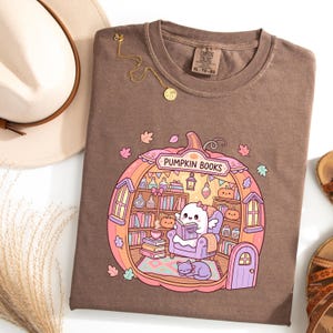 May include: Brown t-shirt with a pumpkin-shaped graphic that reads "Pumpkin Books." The graphic features a ghost reading a book in a cozy library setting. A gold necklace and a hat are also visible.