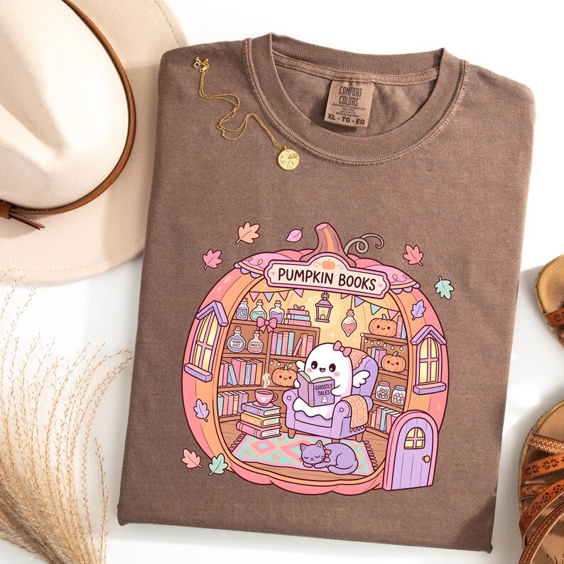 May include: Brown t-shirt with a pumpkin-shaped graphic that reads "Pumpkin Books." The graphic features a ghost reading a book in a cozy library setting. A gold necklace and a hat are also visible.