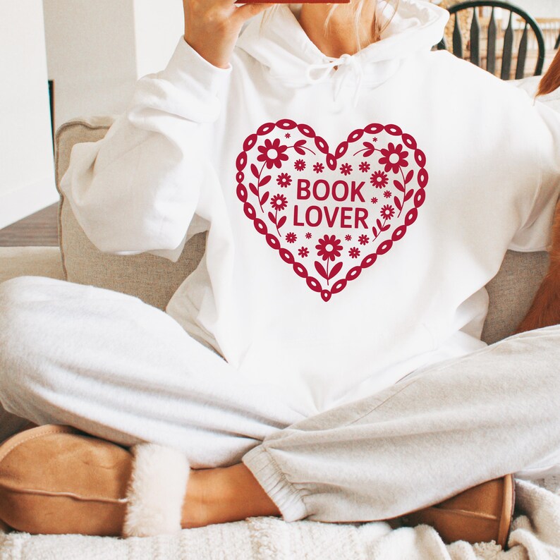 Puede incluir: Sudadera blanca con capucha y dise&ntilde;o de coraz&oacute;n rojo con flores y la frase "BOOK LOVER". La sudadera est&aacute; hecha de un material suave, ideal para los amantes de la lectura.