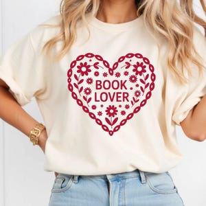 Puede incluir: Camiseta color crema con un dise&ntilde;o en forma de coraz&oacute;n burdeos. El coraz&oacute;n est&aacute; lleno de flores y las palabras "BOOK LOVER". La camiseta tiene mangas cortas y un corte holgado.