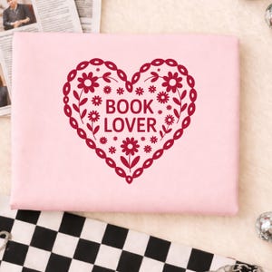 Puede incluir: Una portada de libro rosa claro con un dise&ntilde;o en forma de coraz&oacute;n burdeos. El coraz&oacute;n est&aacute; lleno de flores y hojas, y las palabras "BOOK LOVER" est&aacute;n en el centro. El dise&ntilde;o est&aacute; rodeado por un borde decorativo.