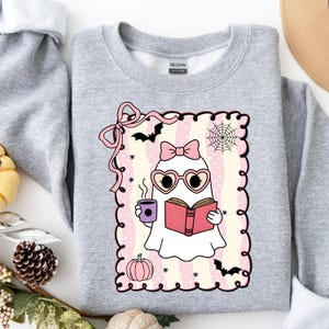 May include: A light gray Gildan sweatshirt featuring a cute ghost illustration. The ghost wears heart-shaped glasses and a bow, holding a book and a coffee cup. The design includes a pink frame, bats, a spiderweb, and a pumpkin.