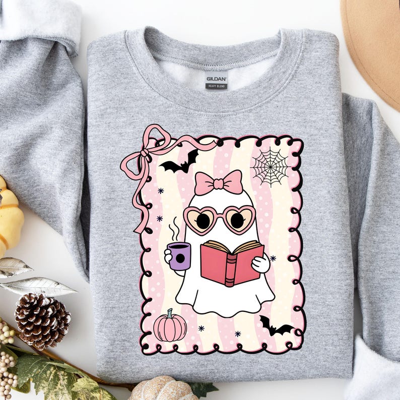 May include: A light gray Gildan sweatshirt featuring a cute ghost illustration. The ghost wears heart-shaped glasses and a bow, holding a book and a coffee cup. The design includes a pink frame, bats, a spiderweb, and a pumpkin.