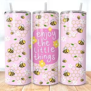 May include: Pink tumbler with a bee design. The tumbler features yellow and black bees, white flowers, and honeycomb patterns. The text "enjoy the little things" is displayed on the front. Includes a silver straw.