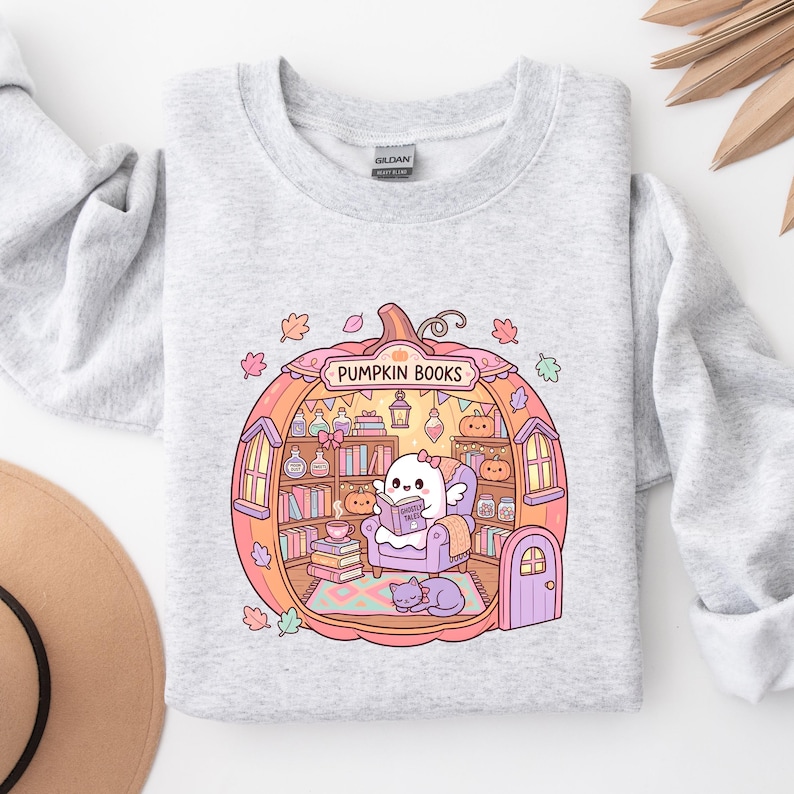 May include: Light gray sweatshirt featuring a whimsical illustration of a ghost reading inside a pumpkin-shaped library. The pumpkin has shelves filled with books and decorative items. The text "PUMPKIN BOOKS" is arched above the pumpkin. A cat is curled up on the floor.