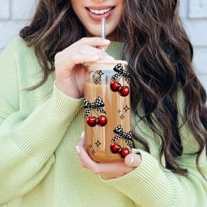 Peut inclure: Un verre transparent avec un couvercle en bambou et une paille en acier inoxydable. Le verre est d&eacute;cor&eacute; de cerises rouges, de n&oelig;uds &agrave; carreaux noirs et blancs et d'&eacute;toiles. Le verre est rempli d'un liquide brun.