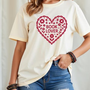 Puede incluir: Camiseta color crema con un dise&ntilde;o de coraz&oacute;n rojo lleno de flores y las palabras "BOOK LOVER". El coraz&oacute;n tiene un borde decorativo. La camiseta tiene mangas cortas y un corte holgado.