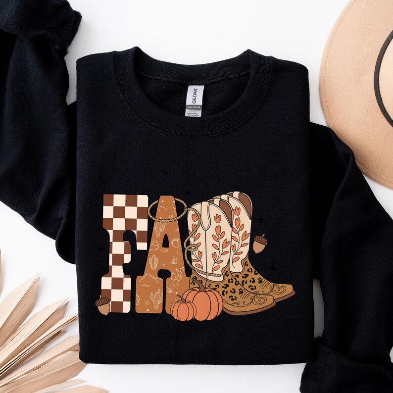 May include: A black long-sleeve sweatshirt featuring a graphic print that spells out the word FALL. The letters incorporate a brown and white checkerboard pattern, a desert cactus motif, and illustrations of cowboy boots, pumpkins, and acorns.