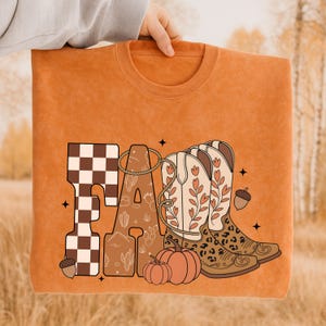 May include: An orange crewneck sweatshirt featuring the word FALL in large letters. The F has a brown and white checkerboard pattern, the A contains cactus line art, and the LL are replaced by two cowboy boots with floral and leopard print.