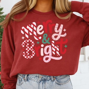 以下が含まれることがあります： お祝いのデザインで「Merry & Bright」の文字が入ったマルーン色のスウェットシャツ。「Merry」は白と赤のストライプ、「&」はティール、「Bright」はピンクと赤で描かれています。スウェットシャツは無地のマルーン色です。