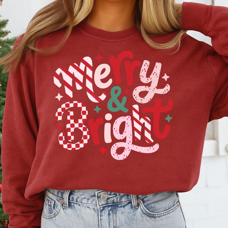 以下が含まれることがあります： お祝いのデザインで「Merry & Bright」の文字が入ったマルーン色のスウェットシャツ。「Merry」は白と赤のストライプ、「&」はティール、「Bright」はピンクと赤で描かれています。スウェットシャツは無地のマルーン色です。