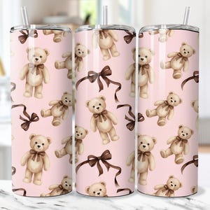 May include: A set of three stainless steel tumblers with a pink background. The tumblers are decorated with a repeating pattern of watercolor teddy bears and brown bows. Each tumbler has a silver lid and a clear straw.