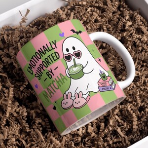 May include: A white ceramic mug featuring a pink and green checkered pattern. The design includes a white ghost wearing heart-shaped sunglasses and bunny slippers, holding a green matcha drink, with the text EMOTIONALLY SUPPORTED BY MATCHA.