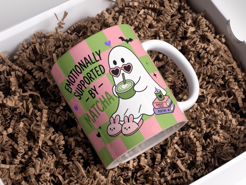 May include: A white ceramic mug featuring a pink and green checkered pattern. The design includes a white ghost wearing heart-shaped sunglasses and bunny slippers, holding a green matcha drink, with the text EMOTIONALLY SUPPORTED BY MATCHA.