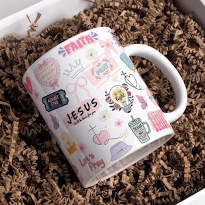 May include: White ceramic mug with a wraparound design featuring religious-themed graphics, including the words "FAITH," "JESUS," and "PRAY." The mug is decorated with pink and pastel-colored illustrations of butterflies, hearts, and flowers. The mug is inside a white box with brown paper filler.