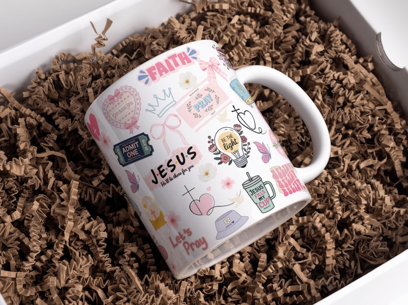 May include: White ceramic mug with a wraparound design featuring religious-themed graphics, including the words "FAITH," "JESUS," and "PRAY." The mug is decorated with pink and pastel-colored illustrations of butterflies, hearts, and flowers. The mug is inside a white box with brown paper filler.