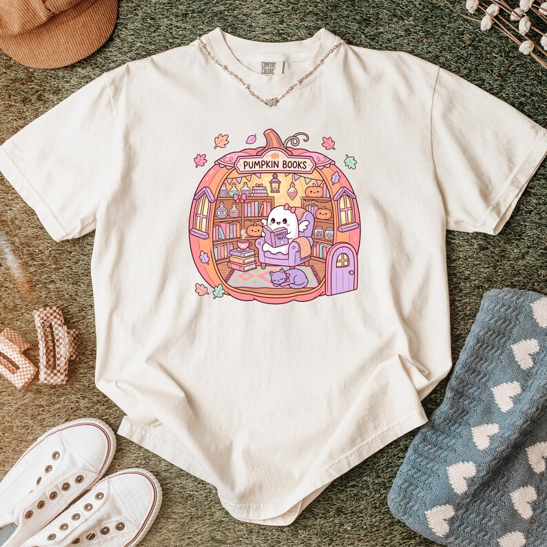 May include: Cream-colored t-shirt featuring a whimsical illustration of a pumpkin-shaped library with the text "PUMPKIN BOOKS". Inside, a cute character reads a book. The design includes bookshelves, a cat, and autumn leaves.