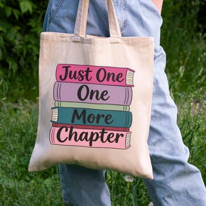 May include: Beige tote bag with the text "Just One More Chapter" stacked over a colorful illustration of books. The books are in shades of pink, purple, teal, and red. The bag has two handles.