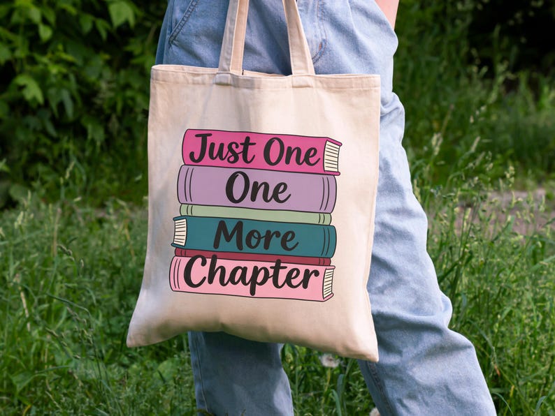 May include: Beige tote bag with the text "Just One More Chapter" stacked over a colorful illustration of books. The books are in shades of pink, purple, teal, and red. The bag has two handles.