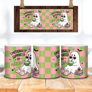 May include: A white ceramic mug featuring a pink and green checkered pattern with a cartoon ghost wearing heart-shaped sunglasses, drinking matcha, and sitting on books. The text reads EMOTIONALLY SUPPORTED BY MATCHA, with small hearts, bats, and slippers.
