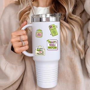 Puede incluir: Un vaso t&eacute;rmico blanco con tapa y asa plateadas, adornado con pegatinas de colores. Las pegatinas presentan texto y gr&aacute;ficos con tem&aacute;tica de matcha. El vaso es sostenido por una persona que lleva un su&eacute;ter beige.