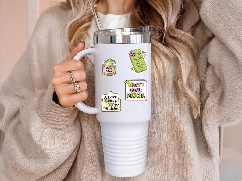 Puede incluir: Un vaso t&eacute;rmico blanco con tapa y asa plateadas, adornado con pegatinas de colores. Las pegatinas presentan texto y gr&aacute;ficos con tem&aacute;tica de matcha. El vaso es sostenido por una persona que lleva un su&eacute;ter beige.