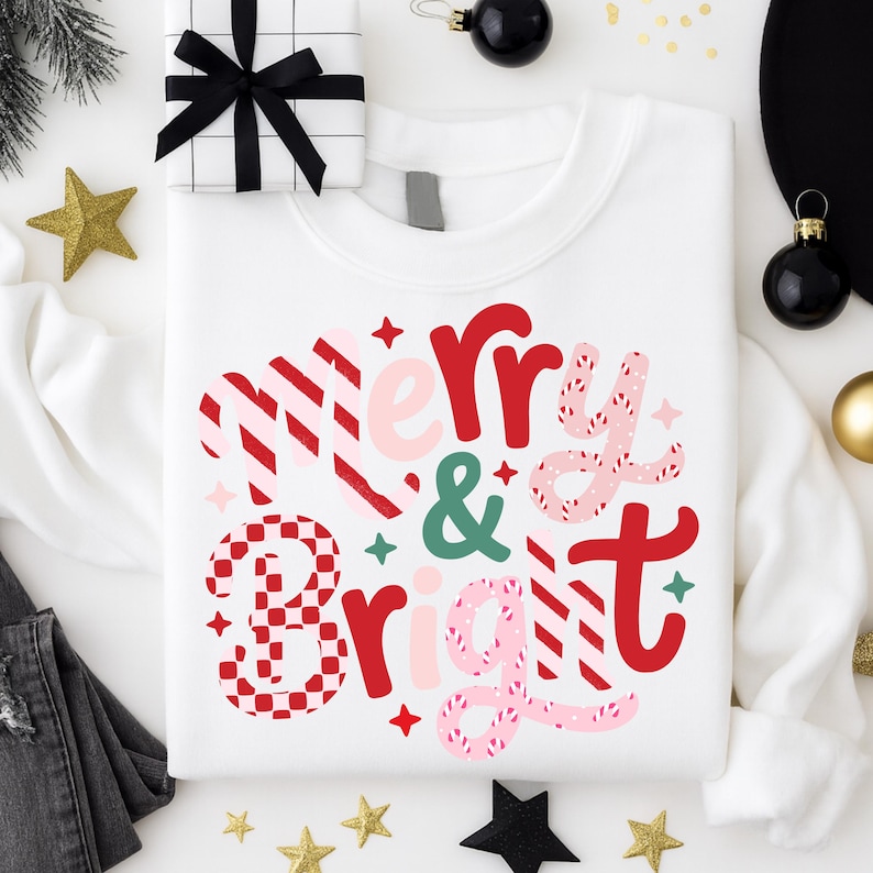 以下が含まれることがあります： 赤、ピンク、緑で「Merry & Bright」と書かれた白いスウェットシャツ。テキストはストライプと市松模様のフェスティバルデザインです。黒いリボンとギフトボックスが背景にあります。