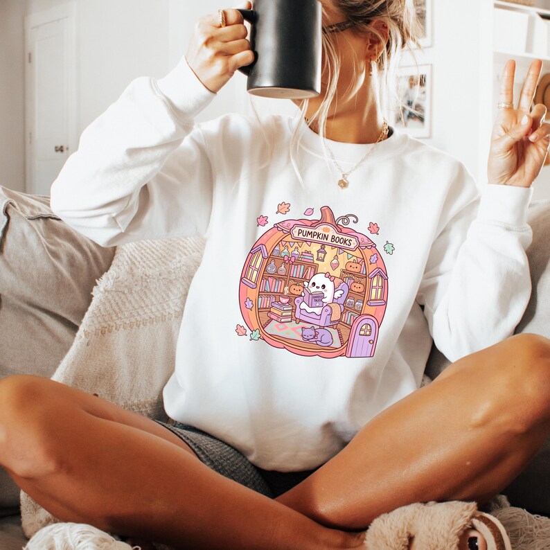 May include: White sweatshirt featuring a colorful graphic of a pumpkin-shaped bookstore with the text "PUMPKIN BOOKS". The illustration includes a ghost reading a book, bookshelves, and a cat. The sweatshirt has long sleeves.