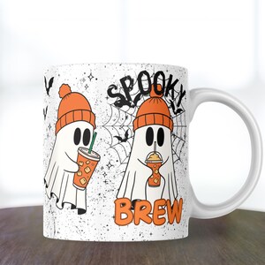 May include: White ceramic mug featuring two cartoon ghosts wearing orange beanies and holding iced drinks. The mug has the words "Spooky Brew" with a spiderweb and bats design, perfect for Halloween.