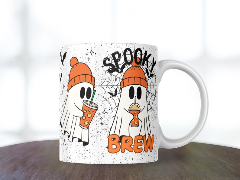 May include: White ceramic mug featuring two cartoon ghosts wearing orange beanies and holding iced drinks. The mug has the words "Spooky Brew" with a spiderweb and bats design, perfect for Halloween.
