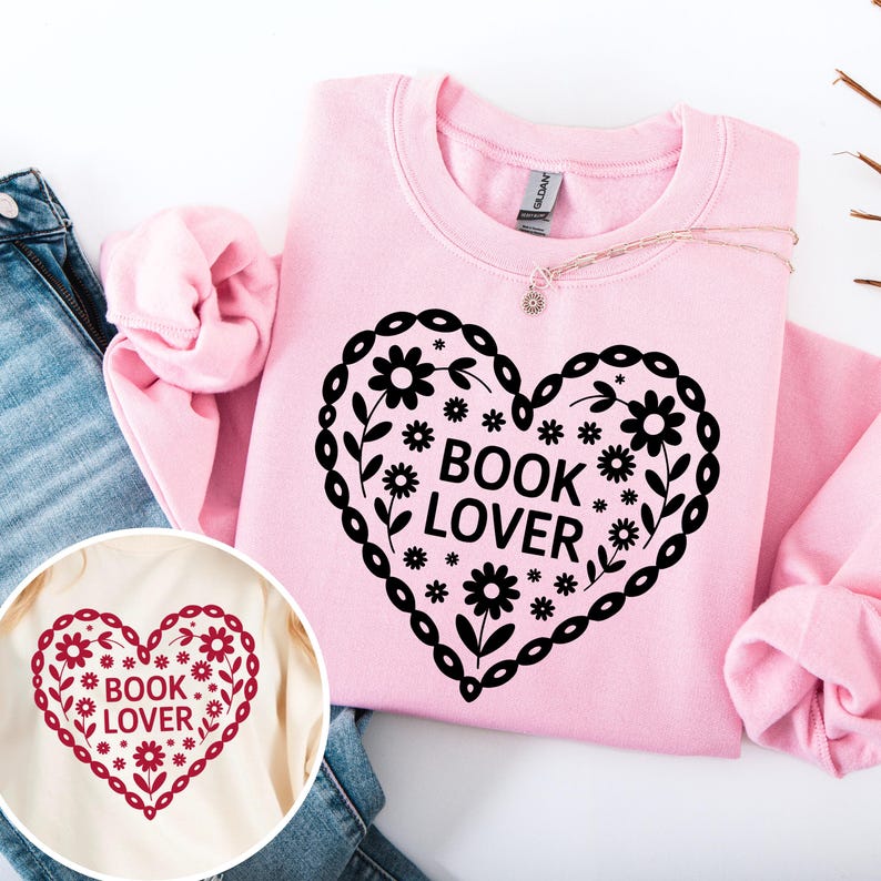 Puede incluir: Sudadera rosa claro con un dise&ntilde;o en forma de coraz&oacute;n negro lleno de flores y la frase "BOOK LOVER". El dise&ntilde;o est&aacute; rodeado por un borde floral. Una versi&oacute;n m&aacute;s peque&ntilde;a del dise&ntilde;o es visible en una camiseta color crema.