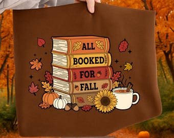 May include: Brown tote bag featuring a stack of books with the words "ALL BOOKED FOR FALL". The design includes fall-themed illustrations like pumpkins, leaves, a sunflower, and a cup of tea. The bag is held by a person.