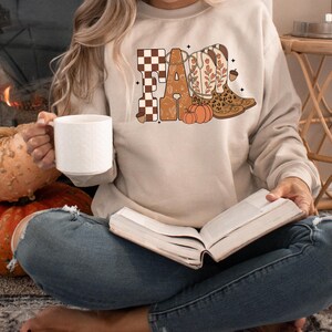 May include: A beige crewneck sweatshirt featuring a graphic print that spells FALL. The letters are decorated with a brown and white checkerboard pattern, floral motifs, and a pair of cowboy boots with leopard print accents and orange pumpkins.