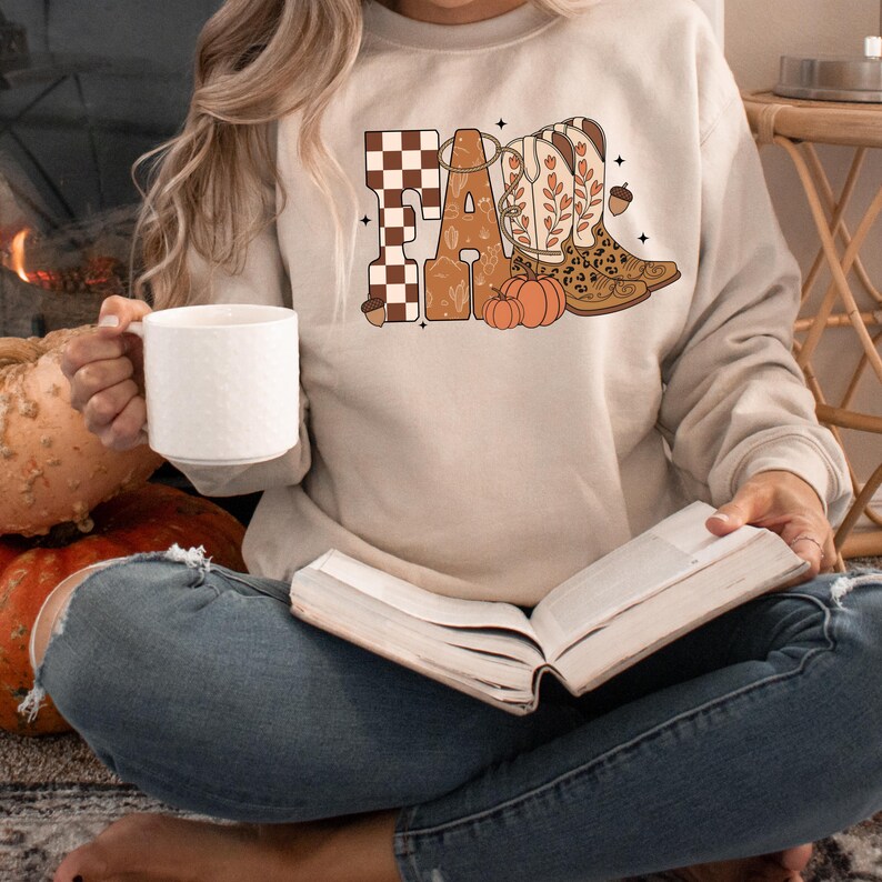 May include: A beige crewneck sweatshirt featuring a graphic print that spells FALL. The letters are decorated with a brown and white checkerboard pattern, floral motifs, and a pair of cowboy boots with leopard print accents and orange pumpkins.