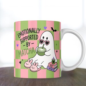 May include: A white ceramic mug featuring a pink and green checkered pattern. The design shows a white ghost wearing heart-shaped sunglasses, drinking a green matcha latte, and wearing bunny slippers. Text reads: EMOTIONALLY SUPPORTED BY MATCHA.