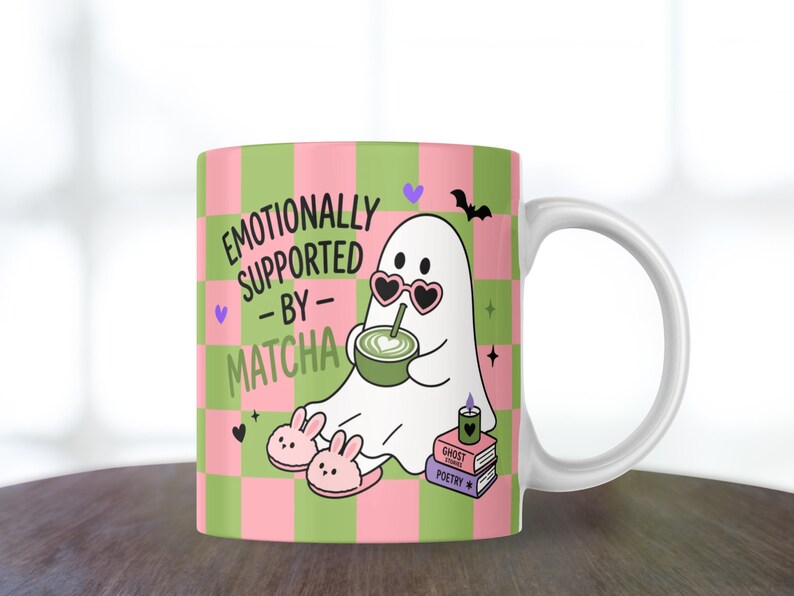 May include: A white ceramic mug featuring a pink and green checkered pattern. The design shows a white ghost wearing heart-shaped sunglasses, drinking a green matcha latte, and wearing bunny slippers. Text reads: EMOTIONALLY SUPPORTED BY MATCHA.