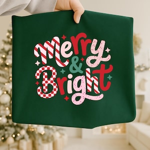 以下が含まれることがあります： 「Merry & Bright」の文字がデザインされた、お祝いの雰囲気の濃い緑色の正方形の布。 赤、白、ピンクの色と星のアクセントが特徴で、ホリデーデコレーションに最適です。 布は人の手で持たれています。