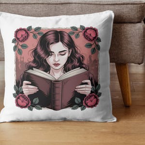 Puede incluir: Coj&iacute;n cuadrado blanco con una ilustraci&oacute;n de una mujer leyendo un libro, enmarcada por rosas rosas y hojas verdes. La mujer tiene cabello oscuro y ondulado y viste una blusa oscura. El fondo es de color rosa p&aacute;lido.