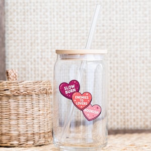 May include: A clear glass tumbler with a bamboo lid and a clear straw. The tumbler has three heart-shaped stickers that read "Slow Burn", "Enemies to Lovers", and "Bookish". The tumbler is designed for cold beverages.