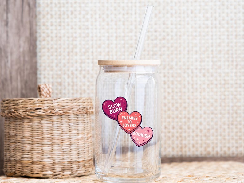 May include: A clear glass tumbler with a bamboo lid and a clear straw. The tumbler has three heart-shaped stickers that read "Slow Burn", "Enemies to Lovers", and "Bookish". The tumbler is designed for cold beverages.