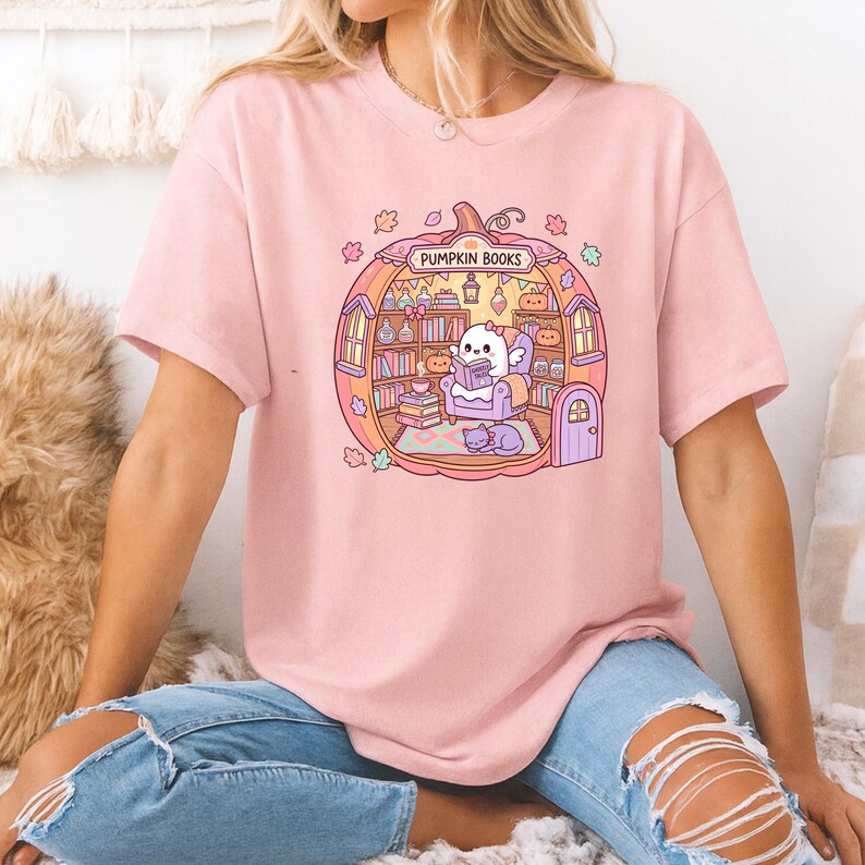 May include: Pink t-shirt featuring a whimsical illustration of a pumpkin-shaped library with the text "PUMPKIN BOOKS". Inside, a cute ghost reads a book. The design includes bookshelves, a cat, and autumn leaves.