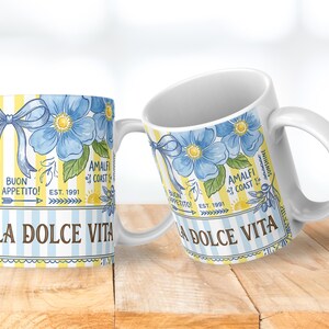 May include: Two white ceramic mugs with a floral design. The design features blue flowers, a blue bow, and the words "LA DOLCE VITA" and "BUON APPETITO!" on a yellow and blue striped background. The mugs are on a wooden surface.