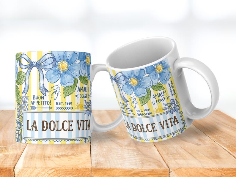 May include: Two white ceramic mugs with a floral design. The design features blue flowers, a blue bow, and the words "LA DOLCE VITA" and "BUON APPETITO!" on a yellow and blue striped background. The mugs are on a wooden surface.
