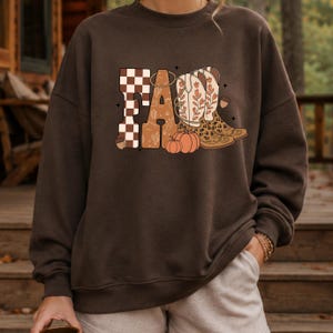 May include: A dark brown crewneck sweatshirt featuring a graphic print on the chest. The design displays the word FALL in large letters, with a checkered pattern, floral cowboy boots, and orange pumpkins integrated into the typography.