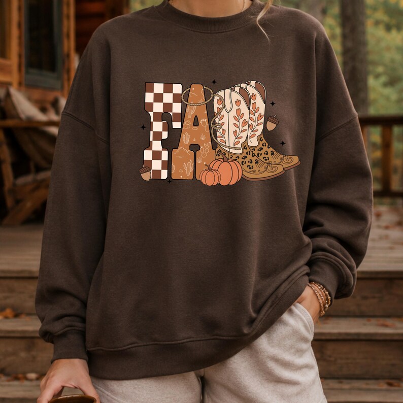 May include: A dark brown crewneck sweatshirt featuring a graphic print on the chest. The design displays the word FALL in large letters, with a checkered pattern, floral cowboy boots, and orange pumpkins integrated into the typography.