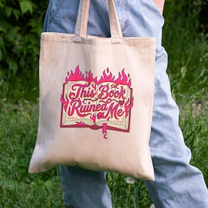 May include: Beige tote bag with the text "This Book Ruined Me" in pink and red, featuring a book engulfed in flames. The bag has two handles and is held by a person wearing blue jeans.