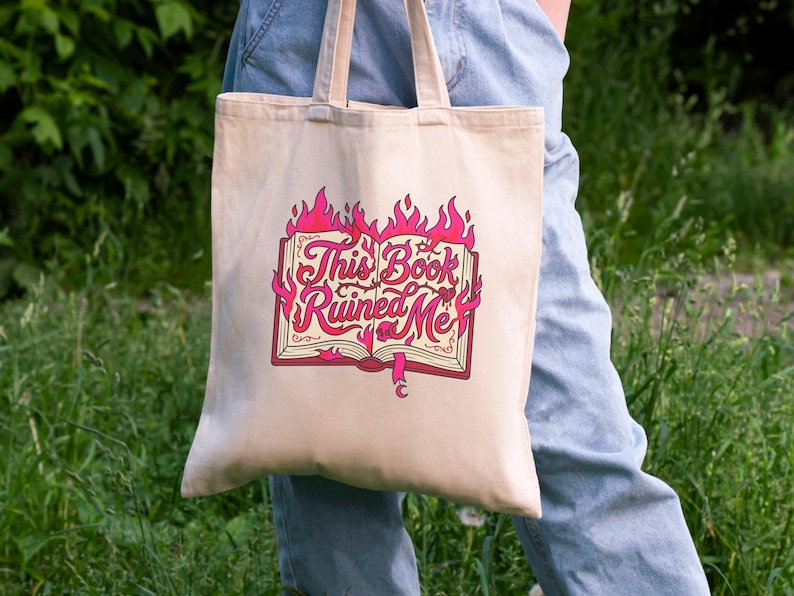May include: Beige tote bag with the text "This Book Ruined Me" in pink and red, featuring a book engulfed in flames. The bag has two handles and is held by a person wearing blue jeans.