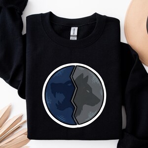 Puede incluir: Sudadera negra con un dise&ntilde;o circular dividido en dos. Un lado muestra un lobo azul aullando, el otro un perfil de lobo gris. El dise&ntilde;o est&aacute; delineado en blanco, creando un contraste llamativo.